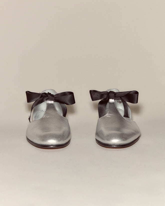 The Theater Shoes - Silver feature T-straps and black ribbon bows, displayed on a simple, light background for a look inspired by the elegance of midcentury dancing shoes.