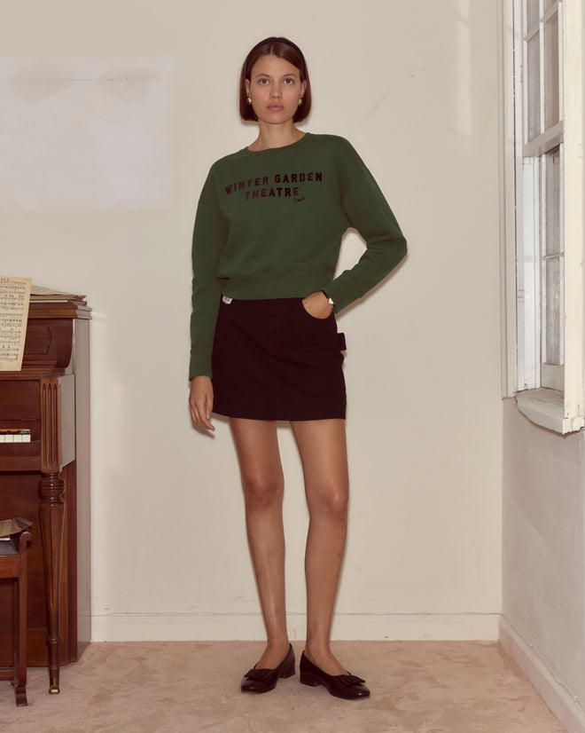 Indoors by a piano, a woman looks at the camera wearing the Theatre Crewneck - Dark Green and a black skirt.