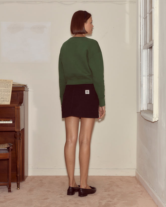 A woman stands indoors facing the wall, wearing a dark green Theatre Crewneck and black skirt, near a piano and window.