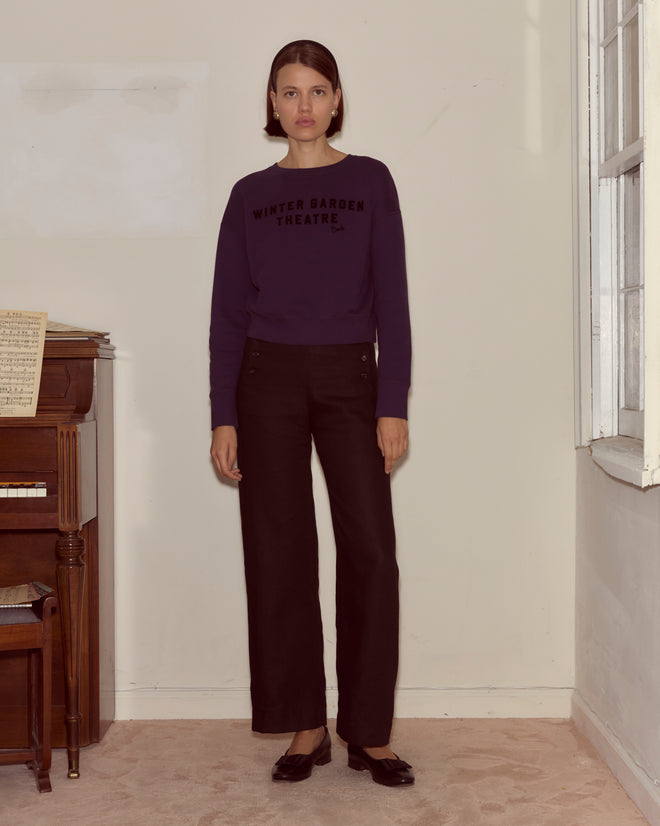 A woman in the Theatre Crewneck - Dark Purple stands indoors by a piano and window, looking at the camera with a timeless Peter Pan-inspired spirit.