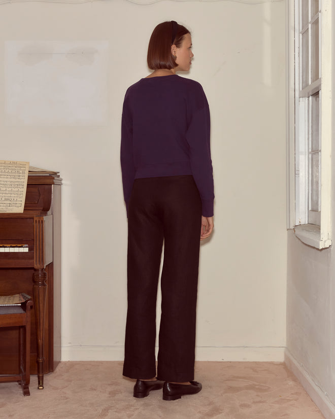A person with short hair stands facing a wall in a room with a piano and window, wearing the Theatre Crewneck in Dark Purple—a look that evokes Peter Pan's quiet charm backstage at the Winter Garden Theatre.