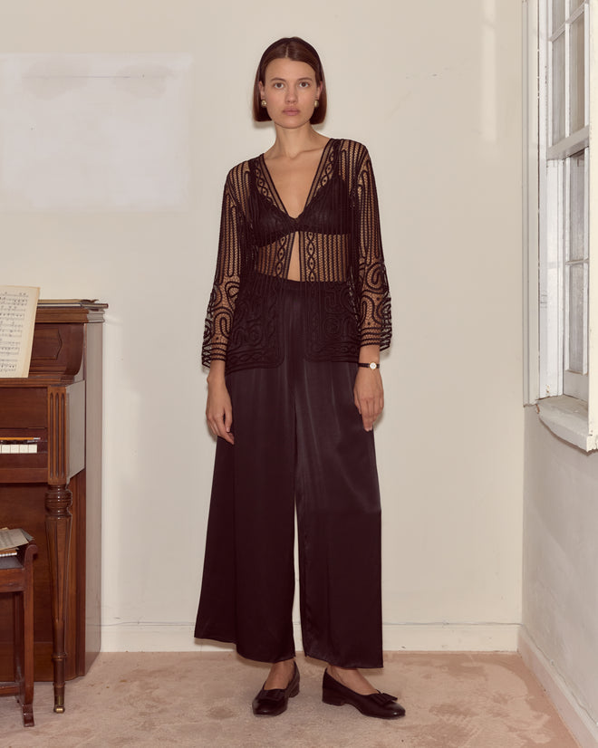 A woman wearing the Threadwork Overshirt, a sheer black lace blouse with delicate hand beadwork, and wide black pants stands by a piano in a minimalist room with white walls.