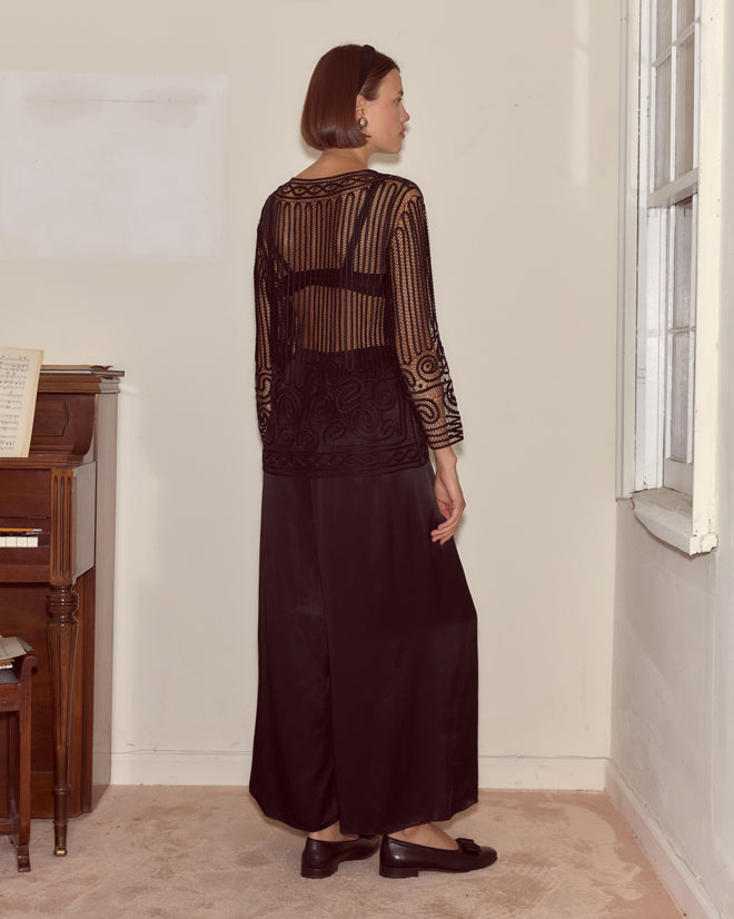 A woman wearing the Threadwork Overshirt, a sheer black top with rhinestone appliqués, and a long black skirt stands by a piano gazing out the window.