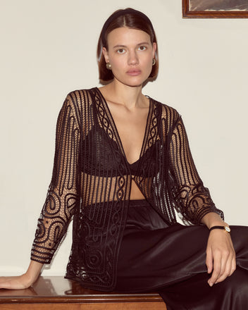 A woman wearing the Threadwork Overshirt, a black sheer embroidered top with delicate hand beadwork, and a skirt sits against a cream wall, looking at the camera.