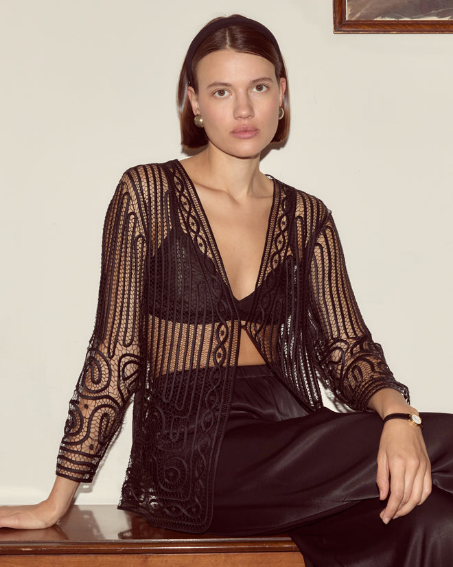 A woman wearing the Threadwork Overshirt, a black sheer embroidered top with delicate hand beadwork, and a skirt sits against a cream wall, looking at the camera.