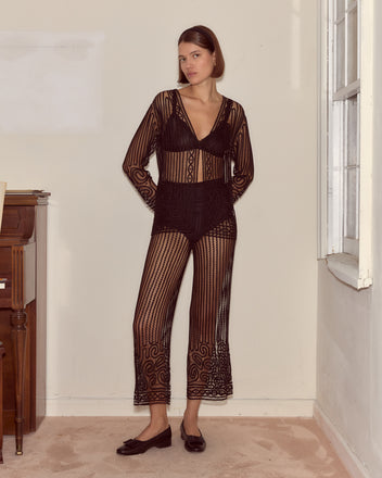 A woman stands indoors near a window, hands in pockets, wearing Threadwork Pants and looking at the camera.