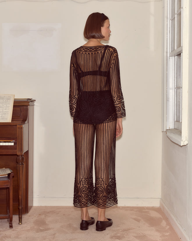 A woman stands facing away in a plain room by a piano and window, wearing Threadwork Pants crafted from sheer black Battenburg lace.