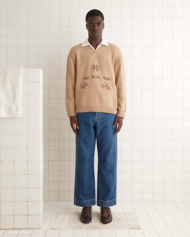 In a tiled room, a person stands wearing the Tic Tac Toe Pullover, an intarsia knit sweater made from soft alpaca wool, featuring a playful Tic Tac Toe pattern. It's stylishly paired with a crisp white collared shirt, blue jeans, and brown shoes.