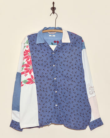 The Tigerseye Napkin Shirt - L/XL is a unique button-up made from vintage napkin fabric, featuring colorful patchwork with floral and animal prints, shown hanging on a hook against a plain wall.