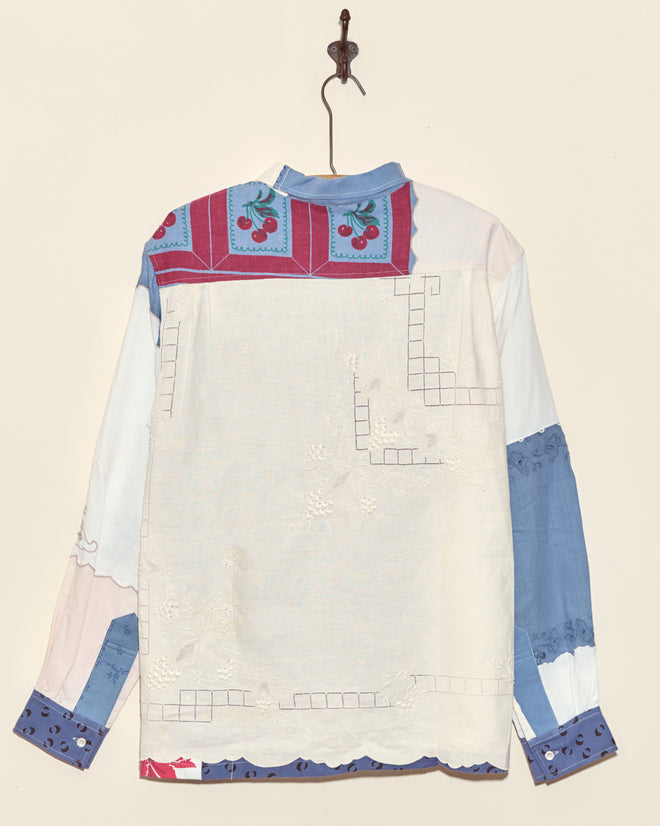 The Tigerseye Napkin Shirt - L/XL, uniquely crafted in New York from blue, white, and pink fabrics with embroidered cherry and floral accents, hangs on a hook.