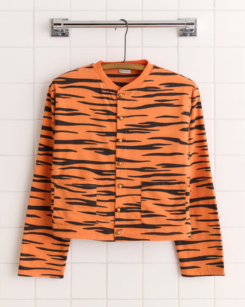 The Tiger Stripe Cardigan, featuring orange and black stripes and brass baseball buttons, hangs on a hook against a tiled wall.