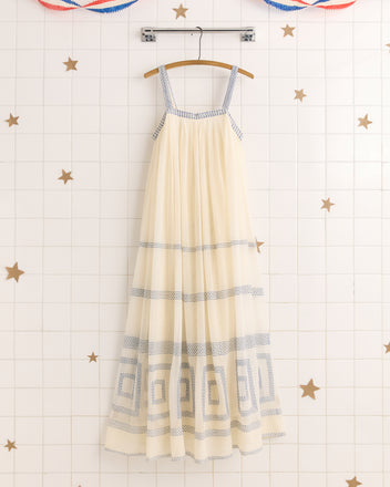 The Tile Stitch Dress, a cream maxi with geometric blue embroidery and blue trim, hangs on a wooden hanger against a white tiled wall adorned with gold stars.