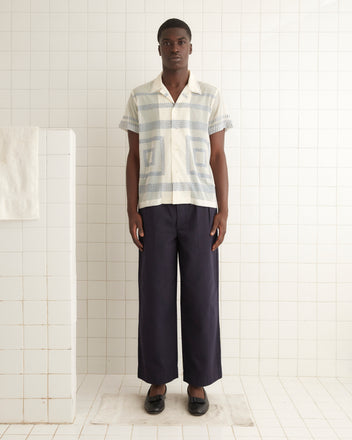 A person stands in a tiled room wearing a Tile Stitch Short Sleeve Shirt with geometric blue embroidery, dark wide-leg pants, and black shoes. The expression is neutral, and the posture is straight.