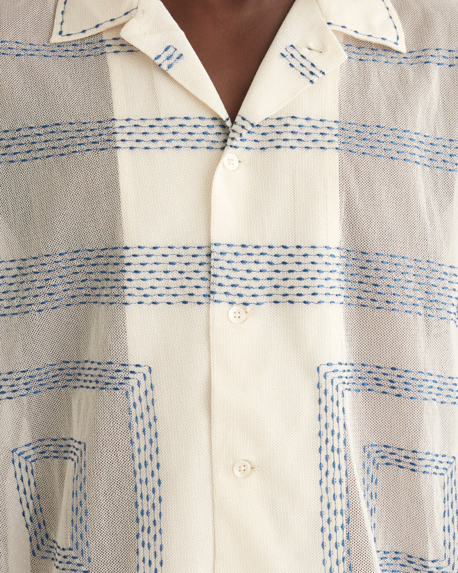 A person stands in a tiled room wearing a Tile Stitch Short Sleeve Shirt with geometric blue embroidery, dark wide-leg pants, and black shoes. The expression is neutral, and the posture is straight.