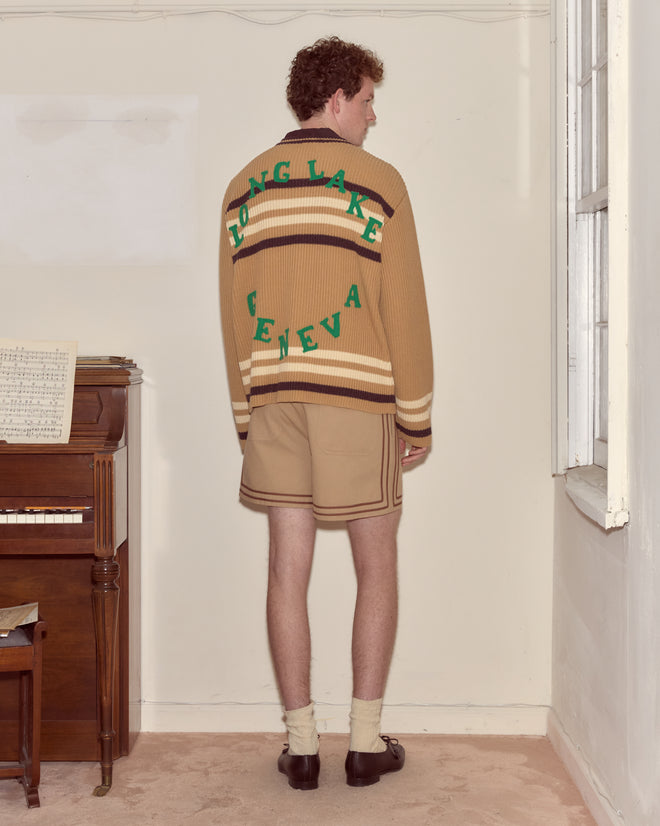 A person stands indoors facing away, wearing a tan Timber Stripe Cardigan that says "LONG LAKE GENEVA," tan shorts, and dress shoes—a look reminiscent of Poconos summer camp style.