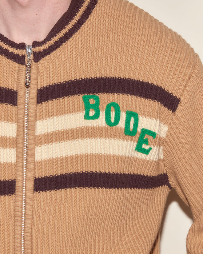 The Timber Stripe Cardigan is a brown knit cardigan with cream stripes and "BODE" in green letters—partially zipped, perfect for cozy camp nights or nostalgic summer adventures.
