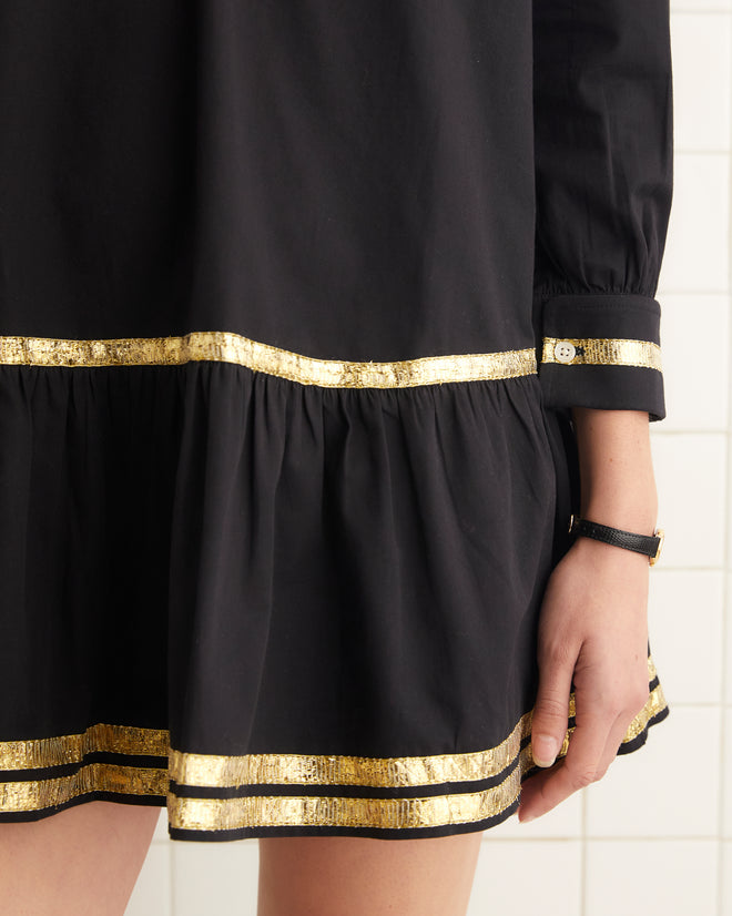 In a white tiled room, a person gracefully stands wearing the Tinsel Tape Dress, featuring a 1930s-inspired design in black cotton twill adorned with striking gold trim.