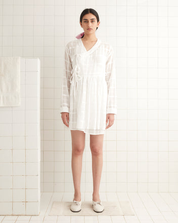 A person in a Tonal Plaid Jama Tunic stands in a pristine, white-tiled bathroom, with a soft cotton towel elegantly hanging on the left.