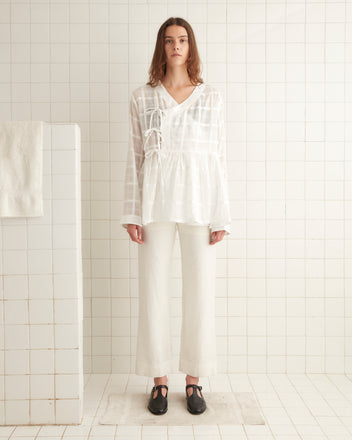 A woman wearing a Tonal Plaid Jama Shirt and pants stands in a tiled room with a towel hanging on a tiled divider.