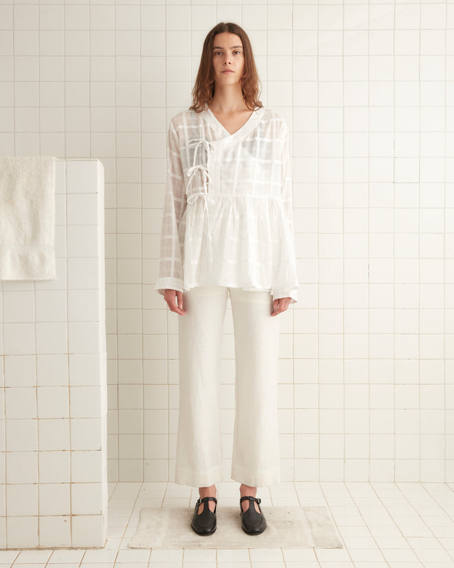 A woman standing in a white tiled room, wearing the Tonal Plaid Jama Shirt and white pants, paired with black shoes.