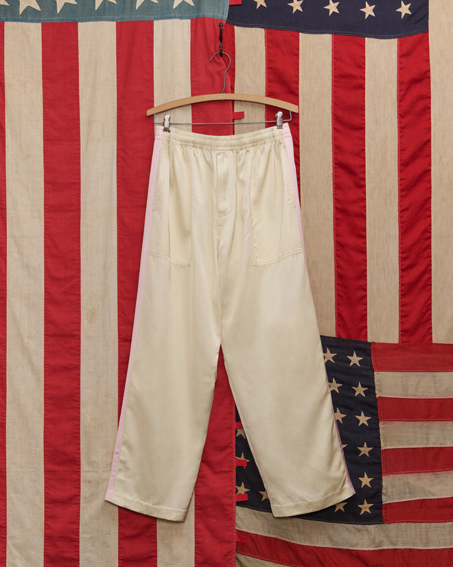 The Satin Track Pants in White/Pink, featuring a subtle side stripe, hang on a wooden hanger against a vintage American flag backdrop.