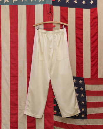The Satin Track Pants in White/Pink, featuring a subtle side stripe, hang on a wooden hanger against a vintage American flag backdrop.
