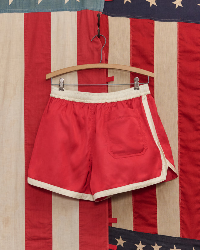 Satin Gym Shorts - Red/White hang on a wooden hanger in front of an American flag background.