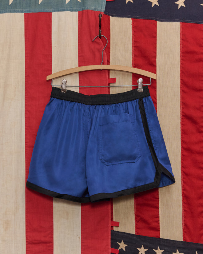 Satin Gym Shorts - Blue/Black hang on a wooden hanger with an American flag background.