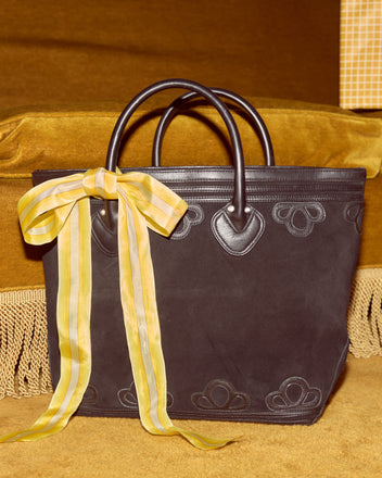 The Trefoil Tote - Black features a boxy silhouette, dark handles, and a yellow striped ribbon bow, displayed on a gold velvet surface.