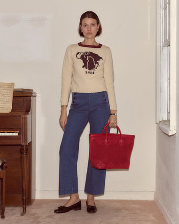 A woman in a boxy animal graphic sweater, blue pants, and holding the Trefoil Tote - Red stands by a piano in a room.