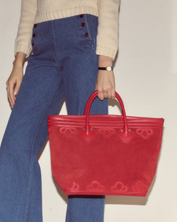 A woman in blue high-waisted jeans carries the Trefoil Tote in red suede, styled with a cream boxy sweater and a watch.
