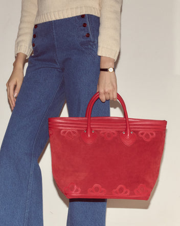 Woman in blue wide-leg jeans carries the Trefoil Tote - Red, paired with a cream sweater and watch, creating a chic look with a boxy silhouette.