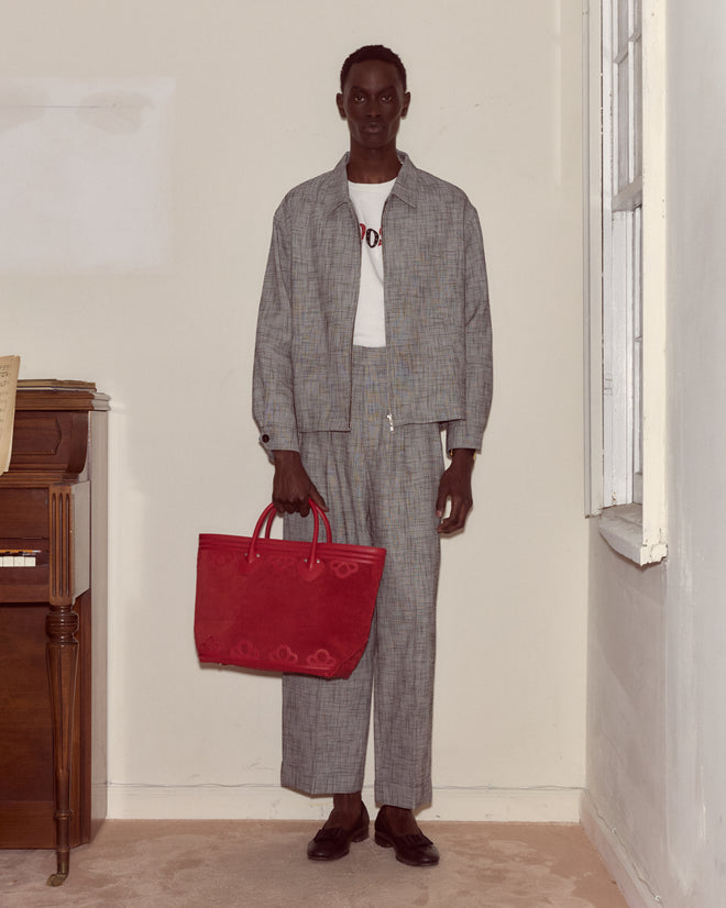 A person in a gray suit holds the Trefoil Tote - Red, a boxy suede bag, while standing indoors beside a piano and window.