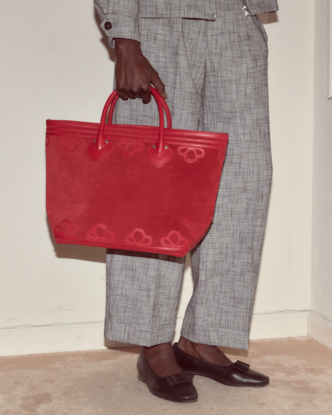 A person in gray suit pants holds the Trefoil Tote - Red, a large suede bag with leather appliqué, and wears black loafers.