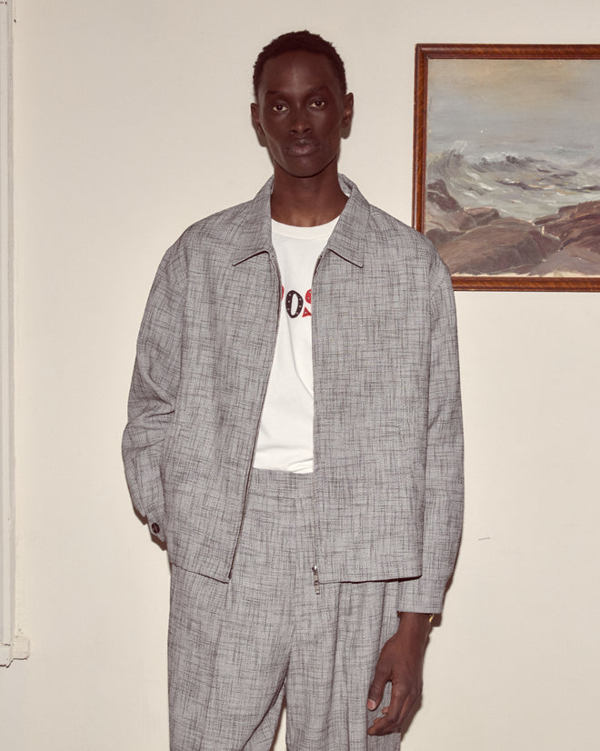 Wearing a Static Weave Ohio Jacket and a white T-shirt, a person in classic suiting stands indoors next to a framed painting on the wall.