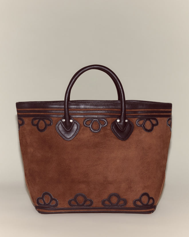 The Trefoil Tote - Brown is a suede bag with a boxy silhouette, dark leather handles, and decorative patterns accenting the top and bottom edges.