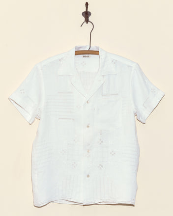 The Trellis Block Short Sleeve Shirt in XS, featuring subtle cutwork embroidery, hangs on a wooden hanger against a beige wall.