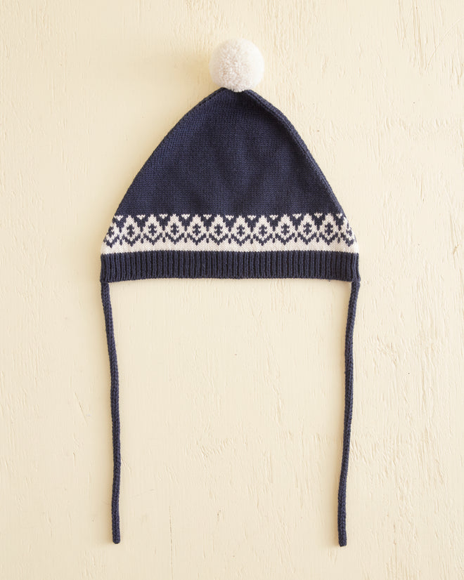 A Navy Bobble Hat, crafted from soft Merino wool and adorned with a playful pom-pom, rests amidst snow and pine branches, embodying the charm of European children's knitwear.