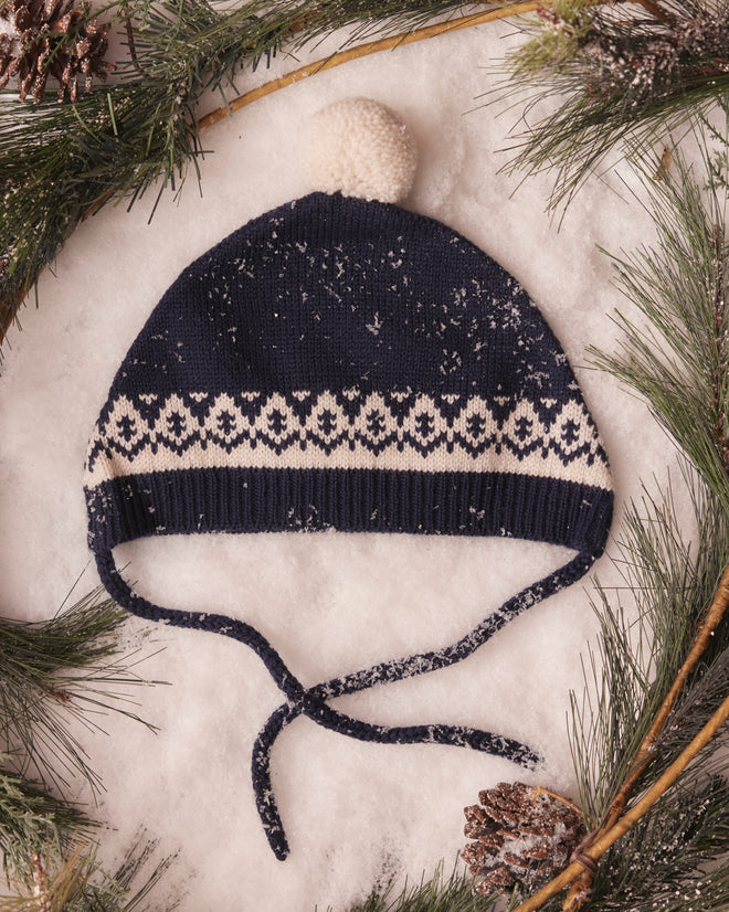 A Navy Bobble Hat, crafted from soft Merino wool and adorned with a playful pom-pom, rests amidst snow and pine branches, embodying the charm of European children's knitwear.