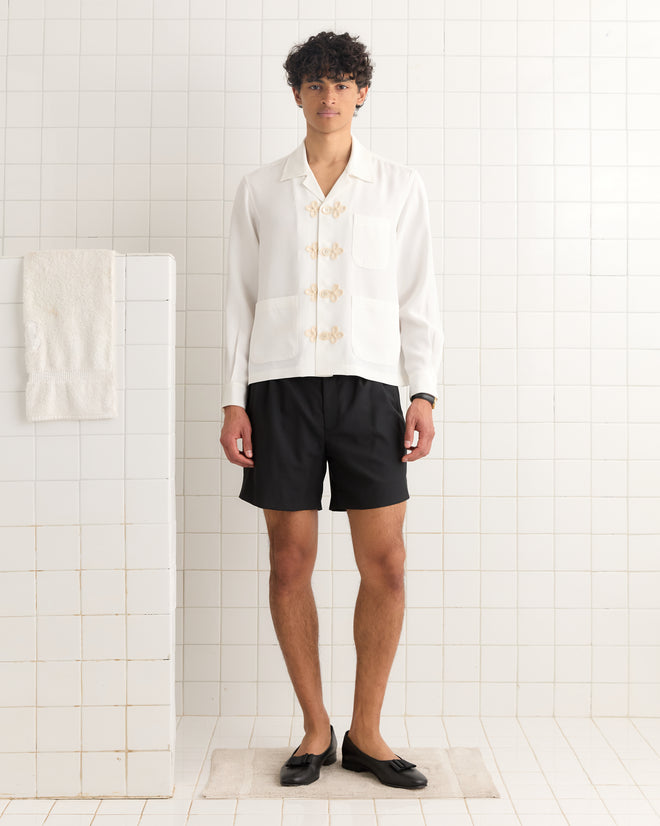 A person stands on a rug in a tiled room, wearing the Trillium Long Sleeve Shirt - Cream, black shorts, and black shoes.