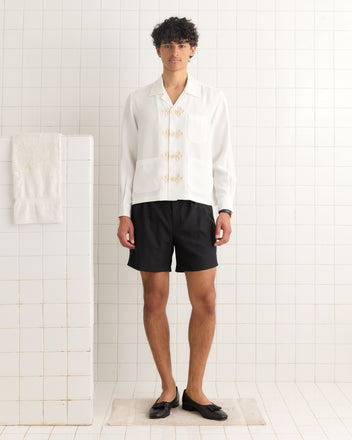 A person stands on a rug in a tiled room, wearing the Trillium Long Sleeve Shirt - Cream, black shorts, and black shoes.