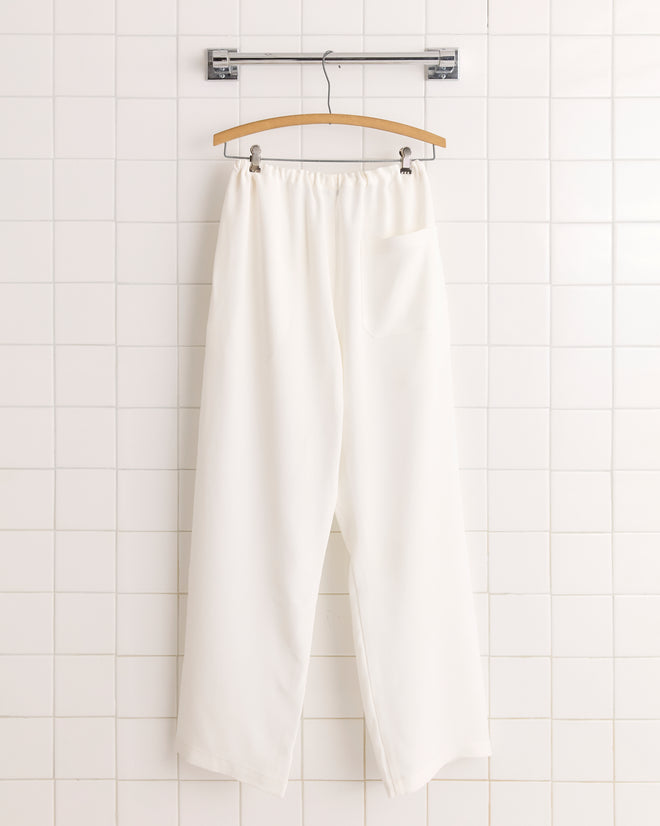 Trillium Pajama Pants - Cream: 1950s-inspired white silk pajama pants featuring an elastic waistband and adjustable drawstring, displayed on a wooden hanger against a tiled wall.