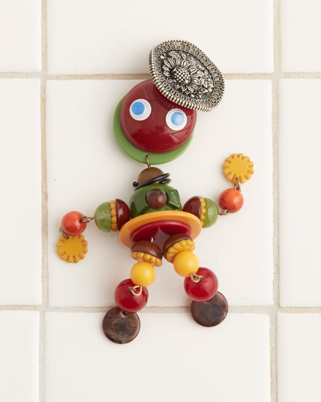 The Trinket Man Brooch, a colorful humanoid figurine crafted from various beads and buttons, evokes the whimsical style of 1920s jewelry. It is attached to a white tiled wall, featuring wide blue eyes, a circular head, and embellishments that suggest arms and legs.