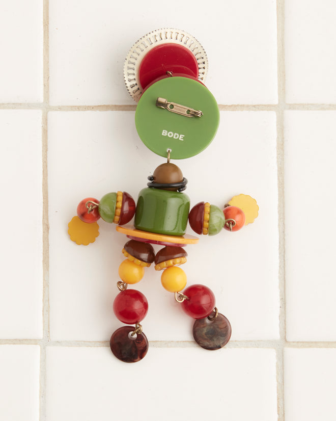 The Trinket Man Brooch, a colorful humanoid figurine crafted from various beads and buttons, evokes the whimsical style of 1920s jewelry. It is attached to a white tiled wall, featuring wide blue eyes, a circular head, and embellishments that suggest arms and legs.