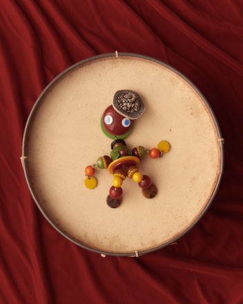 The Trinket Man Brooch, a charming toy figure crafted from beads and buttons, is elegantly displayed on a drum against a vibrant red fabric backdrop, evoking the nostalgic style of vintage accessories.