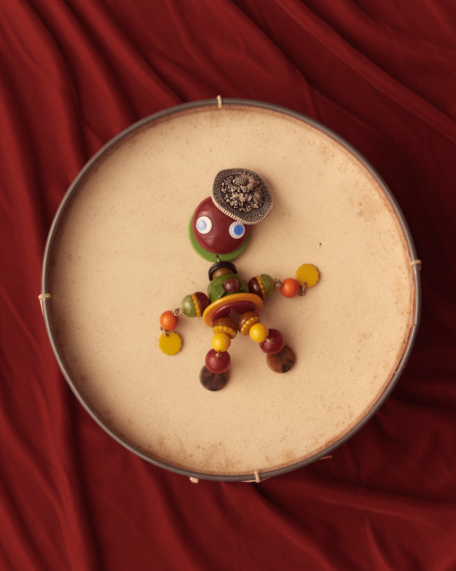 The Trinket Man Brooch, a charming toy figure crafted from beads and buttons, is elegantly displayed on a drum against a vibrant red fabric backdrop, evoking the nostalgic style of vintage accessories.