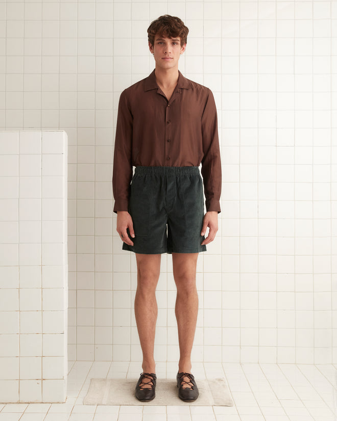 In a tiled room, a person stands wearing a vintage corduroy brown shirt, Tropical Peacock Shorts, and black shoes.