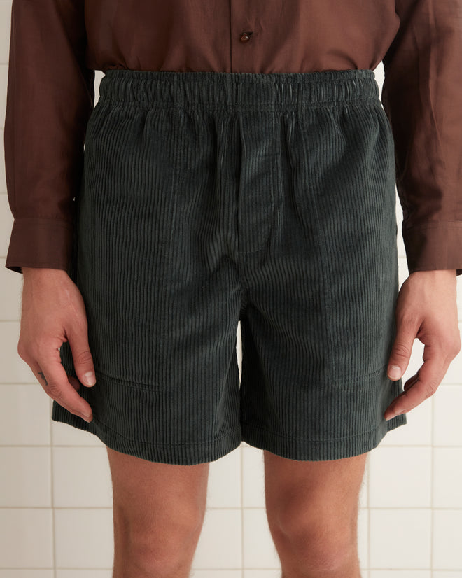 In a tiled room, a person stands wearing a vintage corduroy brown shirt, Tropical Peacock Shorts, and black shoes.