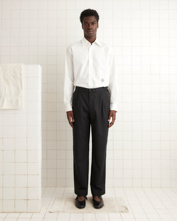 In a tiled room, a person is dressed in a white shirt combined with Tropical Wool Double-Button Trousers, which have a straight-leg design made from lightweight wool, completed by classic black shoes.