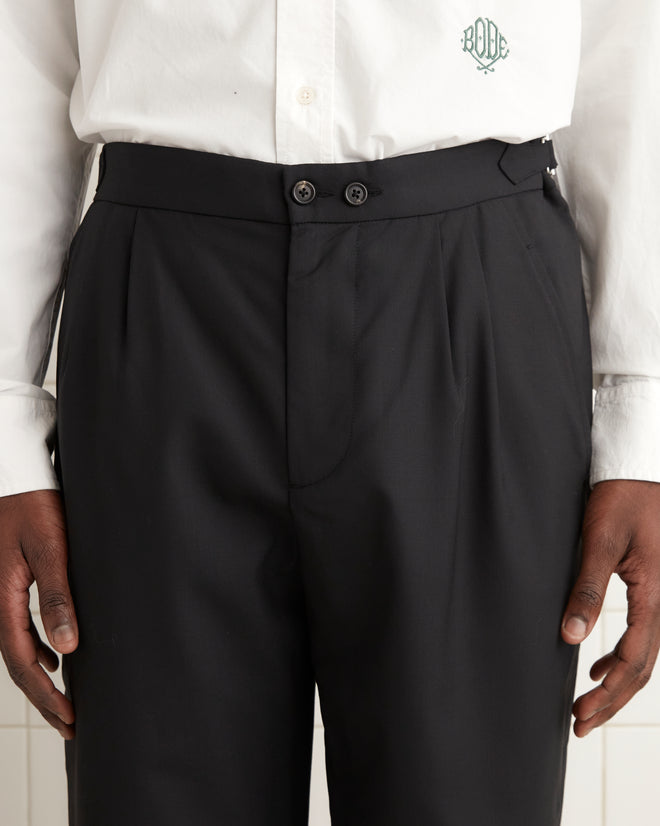 In a tiled room, a person is dressed in a white shirt combined with Tropical Wool Double-Button Trousers, which have a straight-leg design made from lightweight wool, completed by classic black shoes.